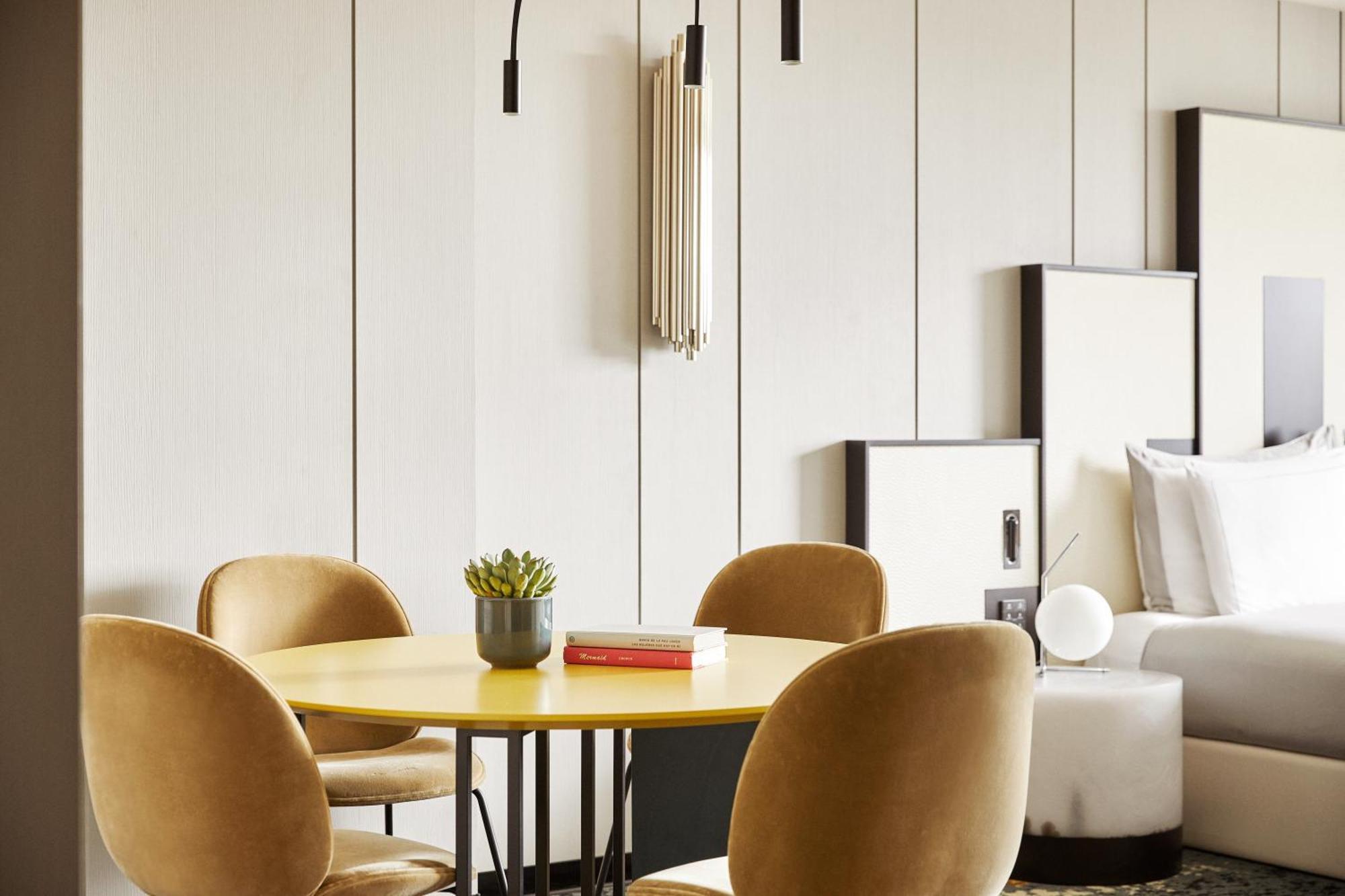 호텔 소피아 바르셀로나 - 인 더 언바운드 컬렉션 바이 하얏트 외부 사진 The photo shows a modern interior setting, featuring a round yellow dining table with four comfortable, curved velvet chairs in a muted color. On the table, there is a small potted plant and a red book. The background features a light-colored wall wi